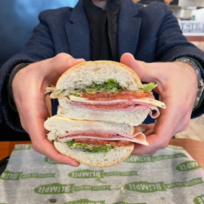 Hands holding stacked Blimpie sandwhich with deli meat, lettuce, cheese, and tomato 