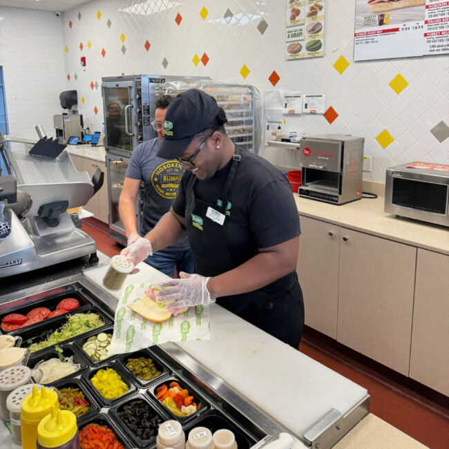Blimpie employee making a deli sandwich with a variety of toppings on display