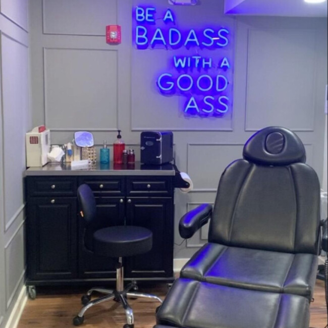 Treatment room with plush black chair and a neon sign on the wall that says "be a badass with a good ass"