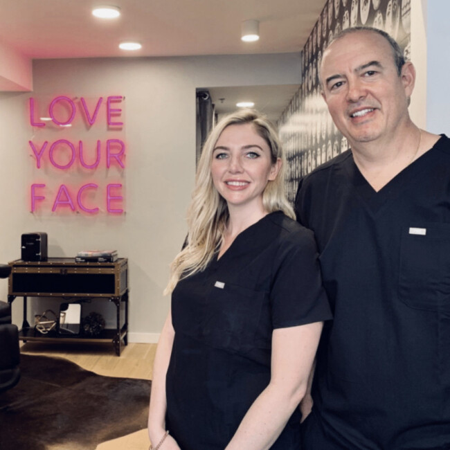 Dr. Oz Fernandez and NP Austin Marie Jacobus standing together in black scrubs in front of a sign that says "love your face"