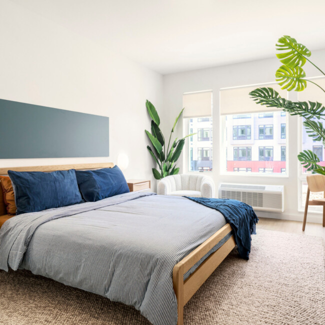 Bedroom inside The Devan with a large bed, gray bedspread, blue pillows, and large window with plants nearby