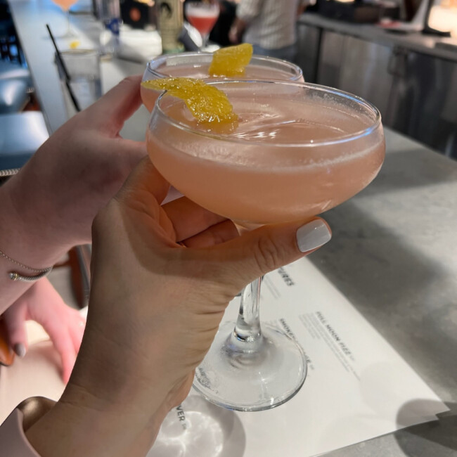 Two hands holding pink cocktails with lemon peels