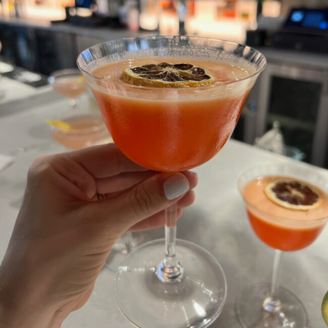 Hand holding an orange cocktail with a dried citrus garnish and another orange cocktail on the bar