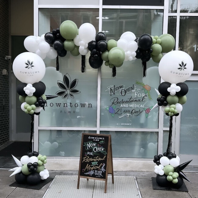 Chalkboard sign and balloon arch with white, green, and black balloons in front of the downtown FLWR storefront