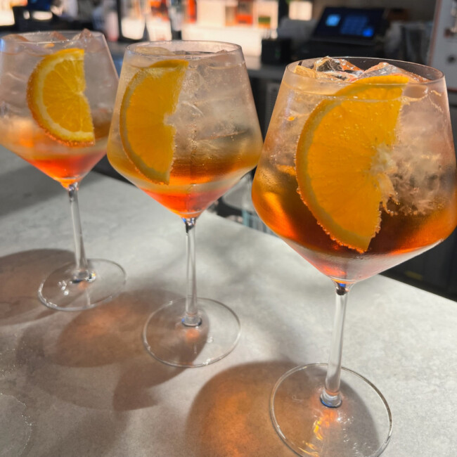 Three Aperol spritz-stlye cocktails in wine glasses on a bar