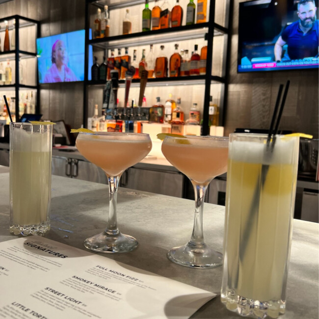 Four cocktails on the bar with two TVs playing in the background