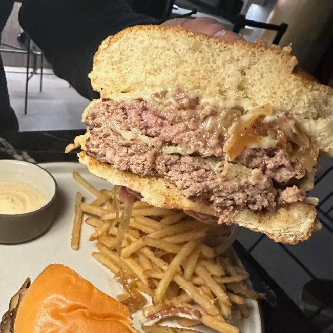 Hand holding half of a burger with a plate of fries and the other half of the burger in background